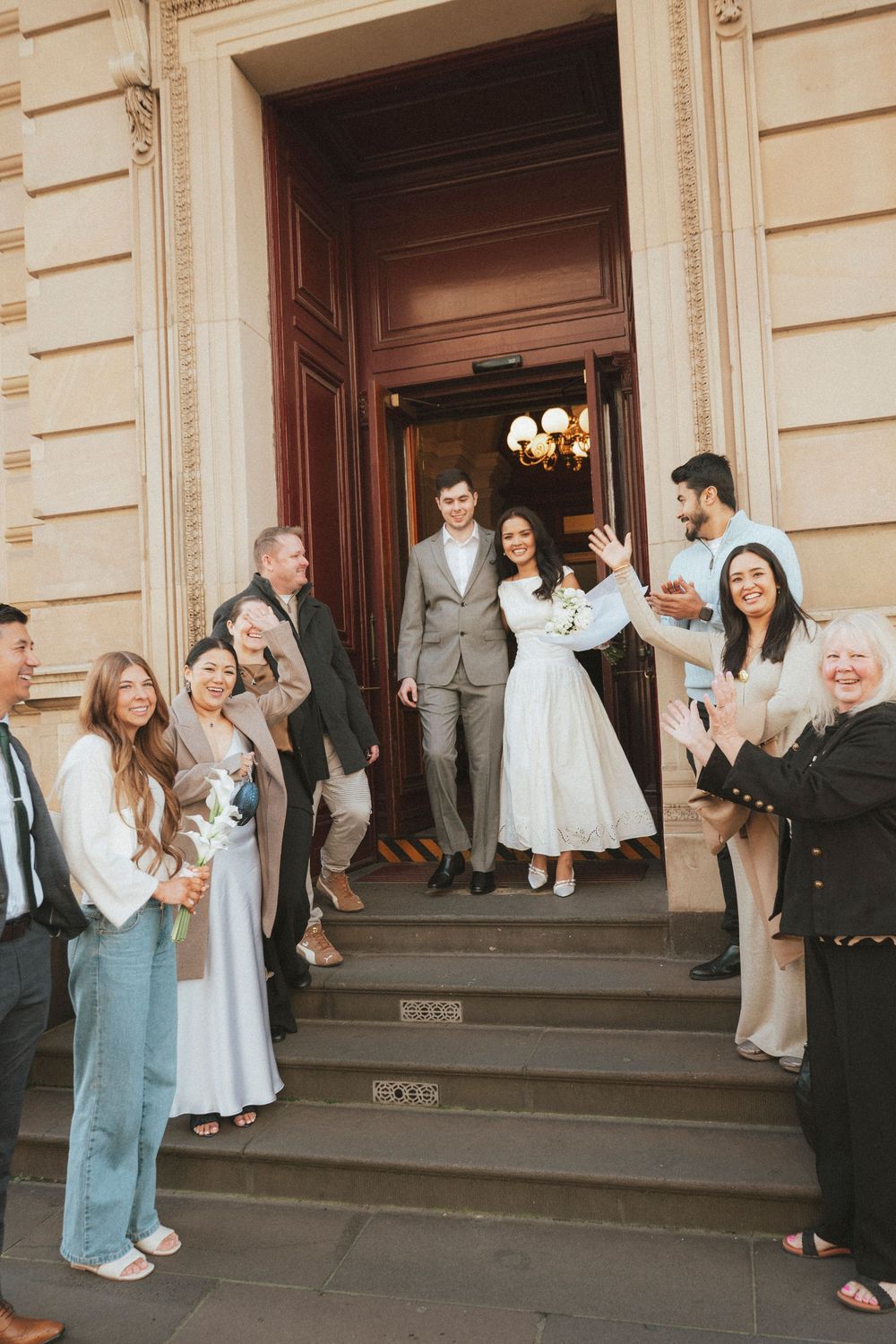 Newlywed couple exits grand building steps surrounded by cheering guests, bride in white dress holding bouquet.