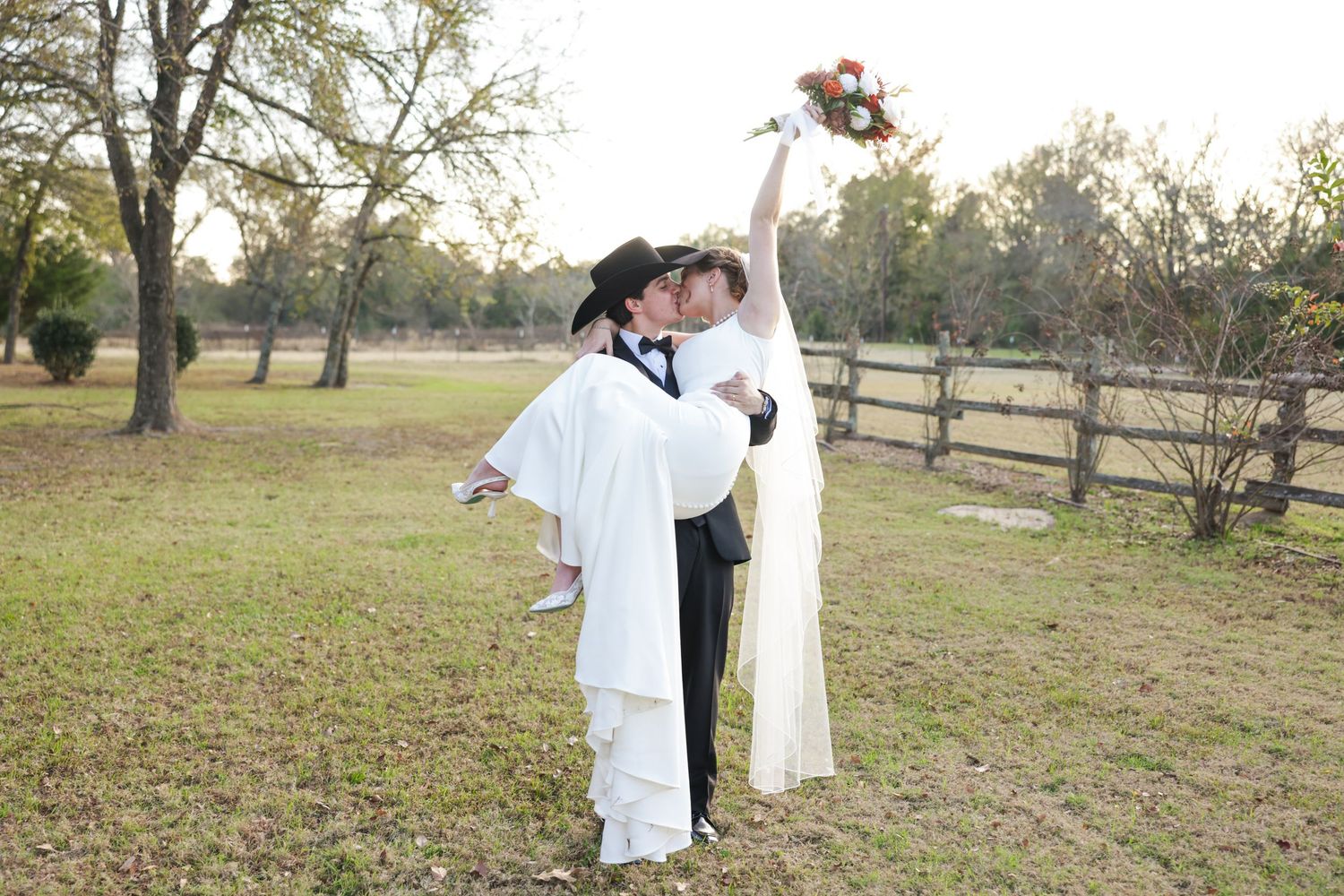 Groom carries bride in white gown outdoors at rustic farm wedding venue, bride raises bouquet joyfully.