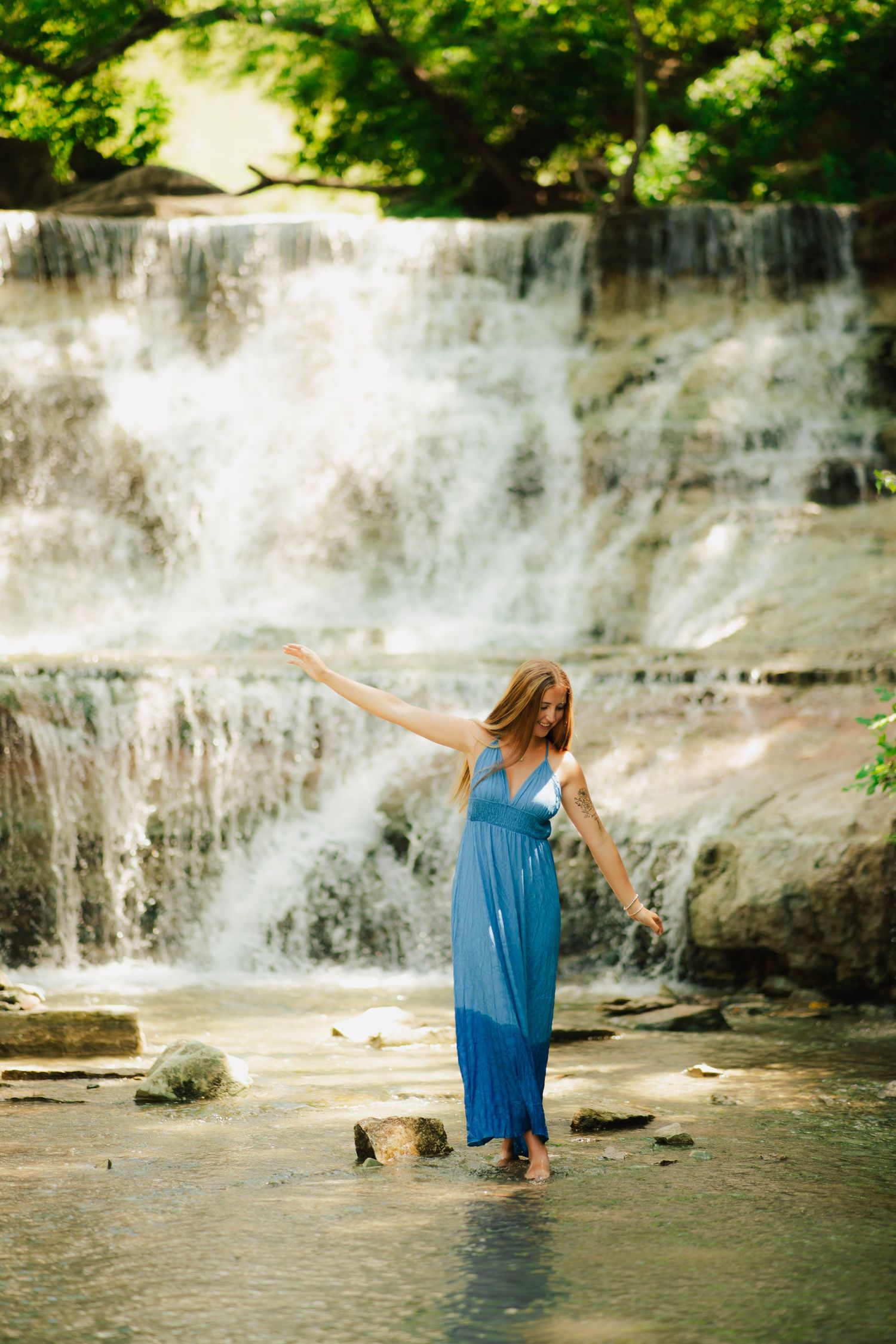 Woman in blue maxi dress standing in shallow water before a cascading waterfall surrounded by lush green trees.