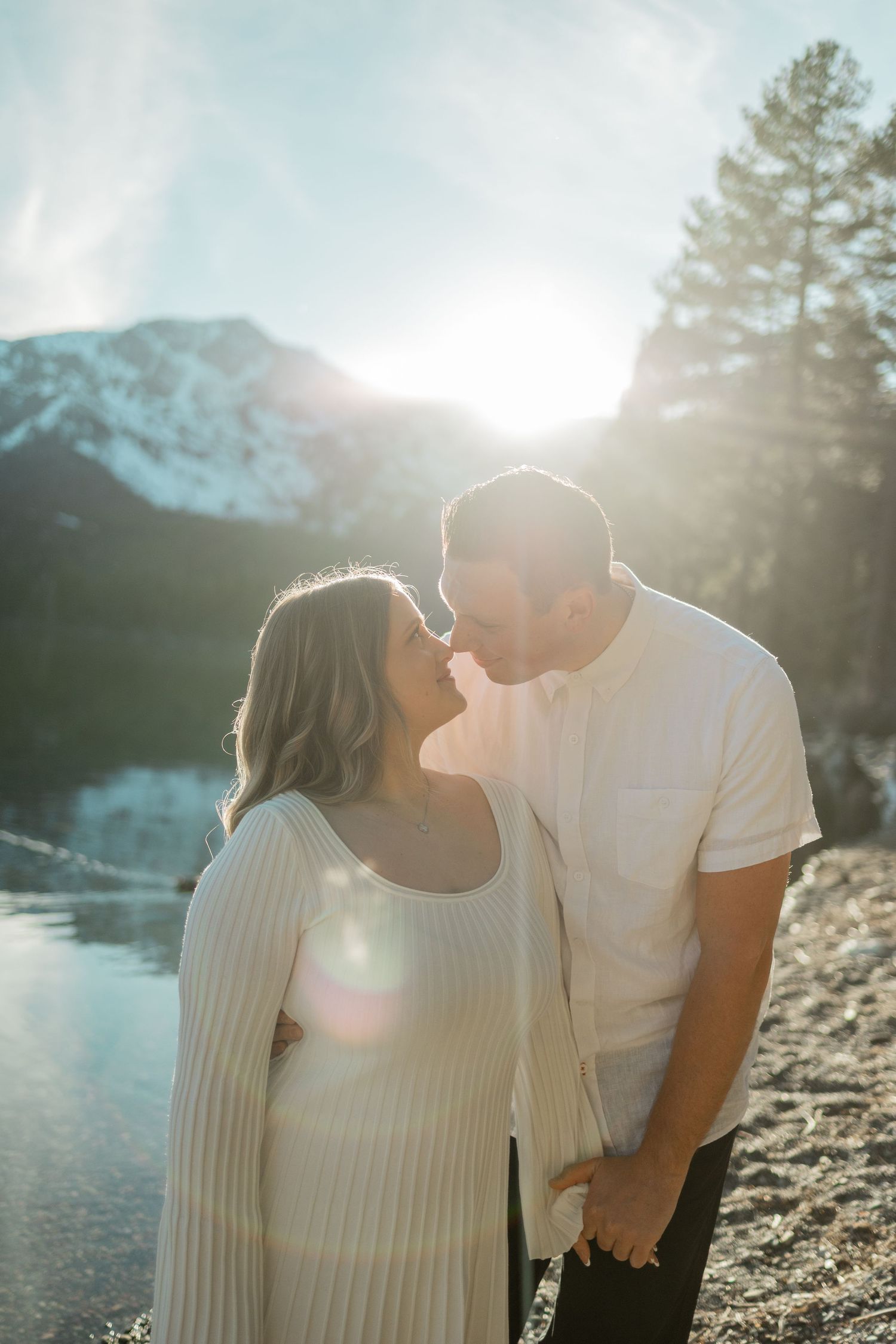 Couple holding hands walking along rocky lakeshore with snow-capped mountains in background on sunny day.