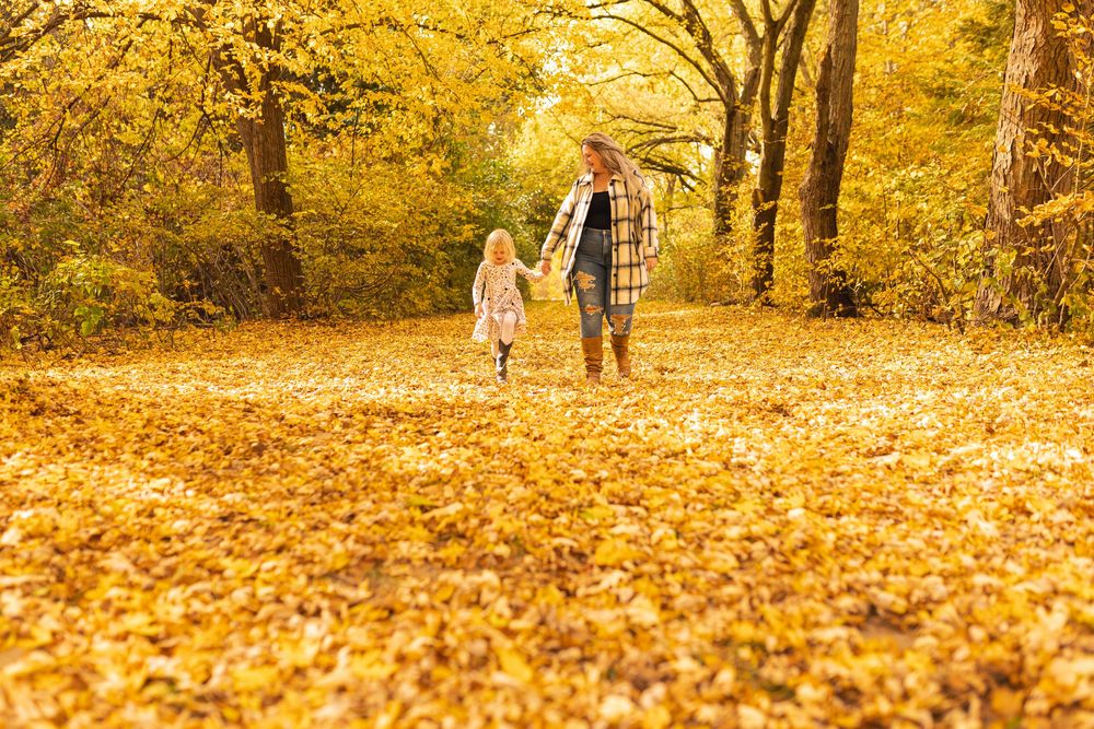 Adult and child walk hand-in-hand through a golden autumn forest path covered in fallen yellow leaves.