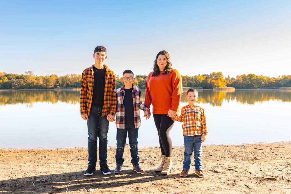 Family of four in coordinating fall outfits posing by a lake on a sunny autumn day.