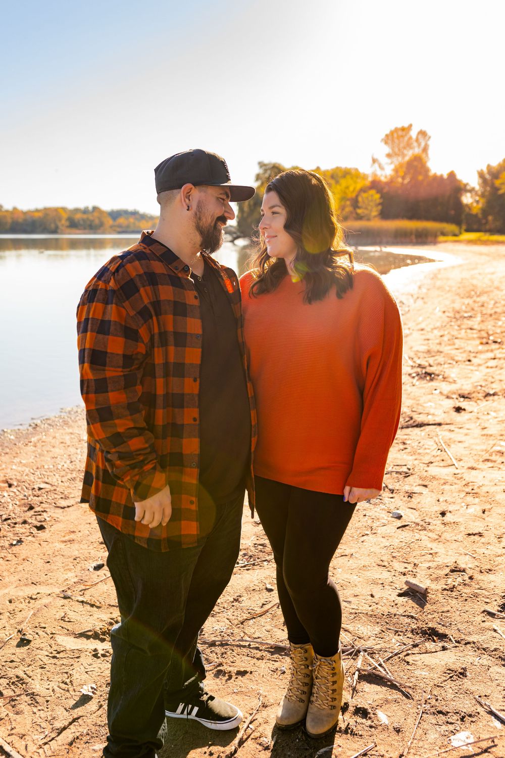 Couple in matching orange outfits smiling at each other by a sunlit lake shore in autumn.