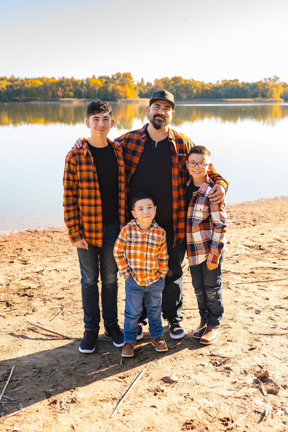 Father and three sons in matching orange plaid shirts pose for a fall family photo on a sandy lakeside shore.