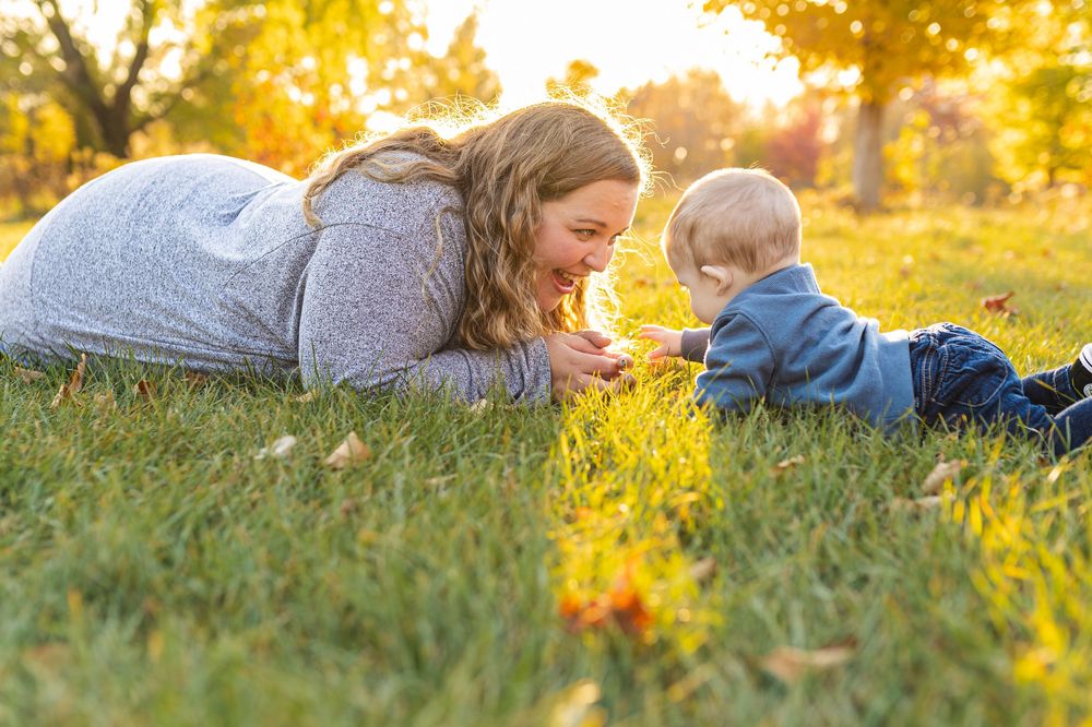 A mother and baby boy lying on green grass in a sunny autumn park, sharing a joyful moment together.