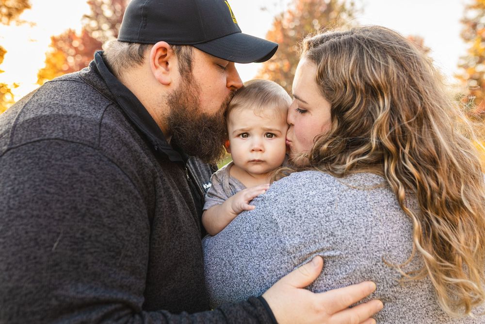 Parents kissing their baby on the cheeks outdoors in autumn, family portrait with fall foliage background.