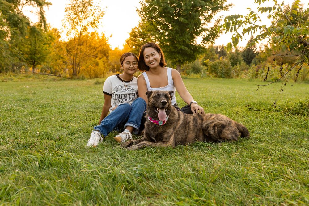 Two young women sit on green grass with a brindle dog at golden hour in a lush park setting.
