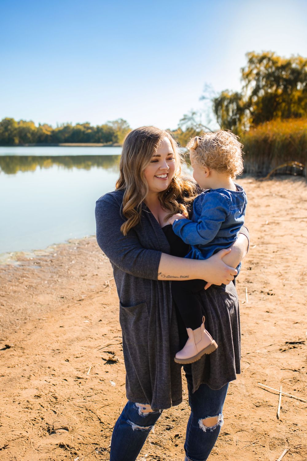 A smiling woman holds a young toddler on a sandy riverbank surrounded by autumn trees and blue sky.