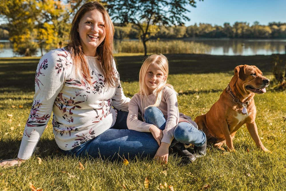 Pregnant woman and young girl sitting on grass with a brown dog near a lake on a sunny autumn day.