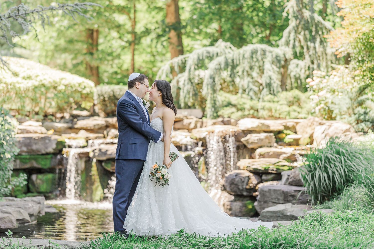 Bride and groom share a romantic kiss in a lush garden with a waterfall backdrop on their wedding day.