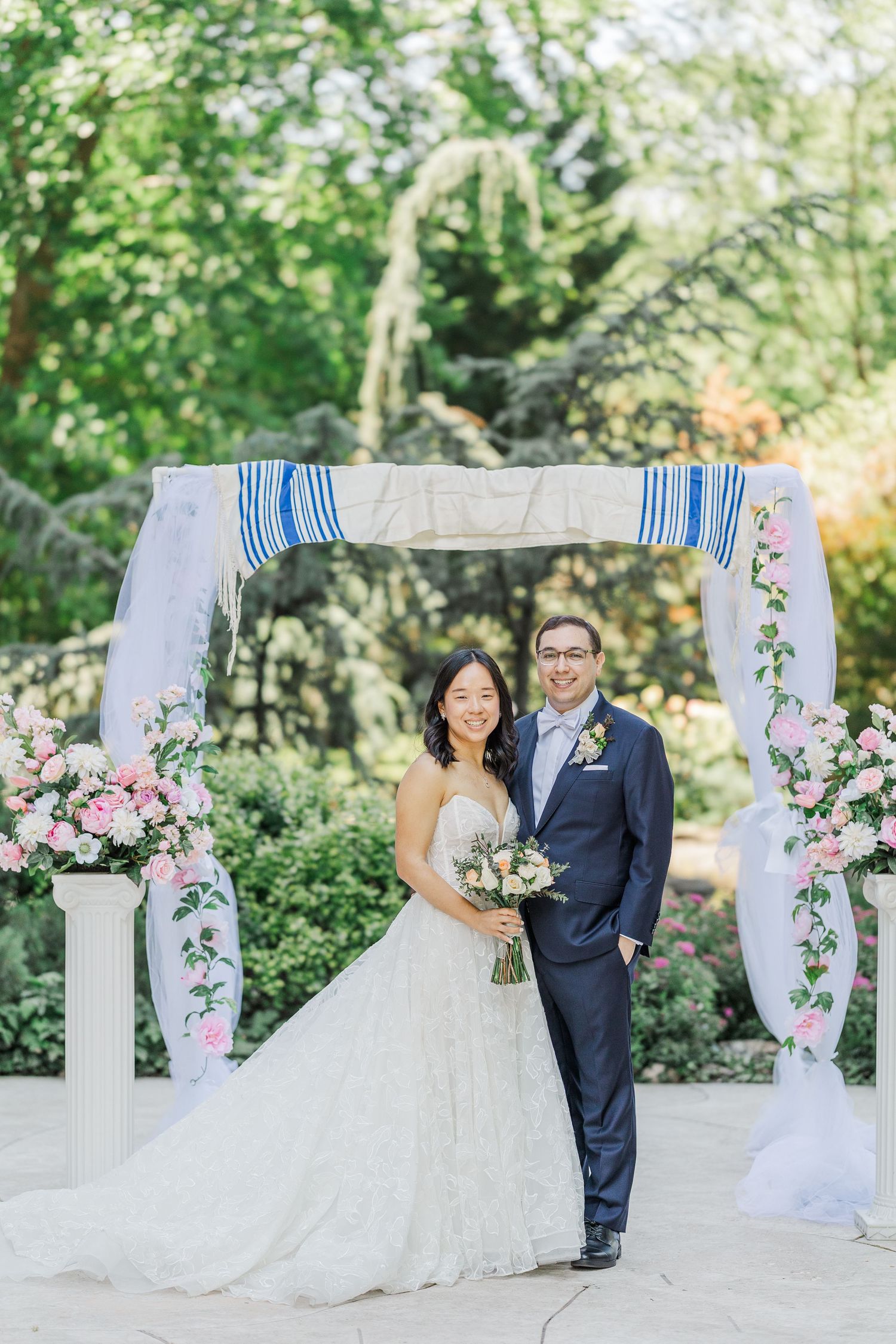 Bride and groom smiling under a floral chuppah with pink roses and white draping in a lush garden wedding ceremony.
