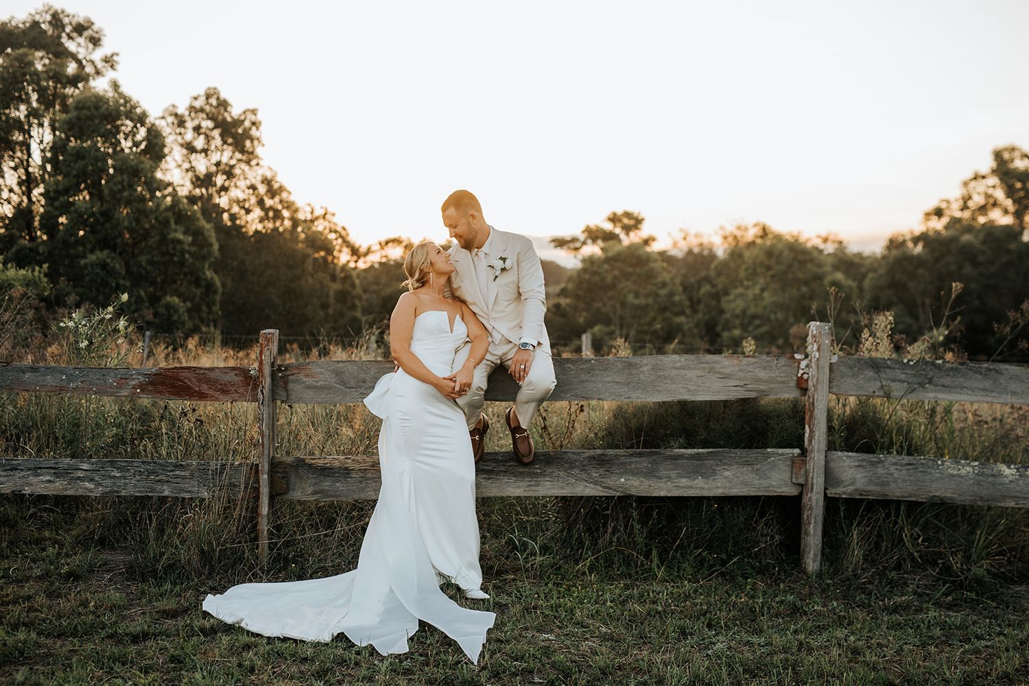Bride and groom pose at golden hour by a rustic wooden fence in a scenic outdoor countryside wedding setting.