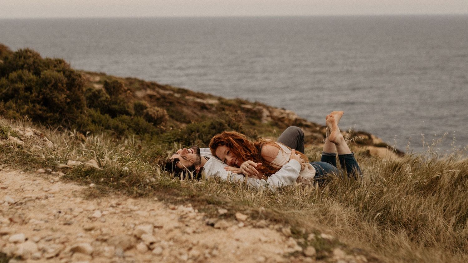 Couple lying on a grassy coastal cliff, sharing a romantic moment with the ocean in the background.