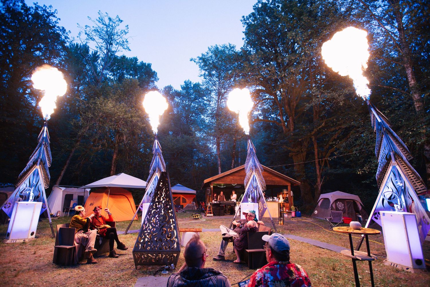 Outdoor festival campsite at dusk with fire towers blazing, decorated tents, and guests gathered among trees.
