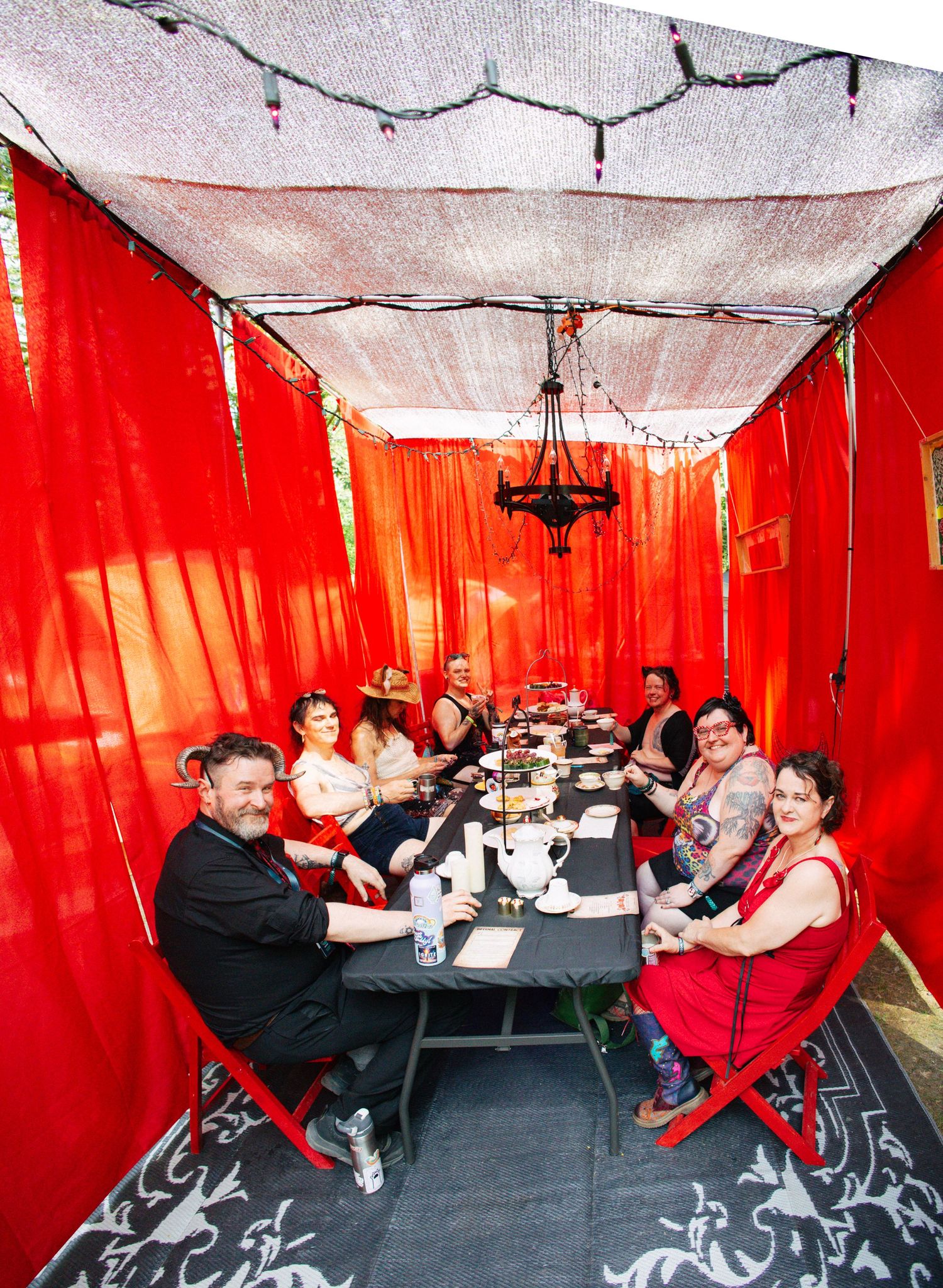 Group dining under red draped tent with chandelier, black table, red chairs, string lights overhead.