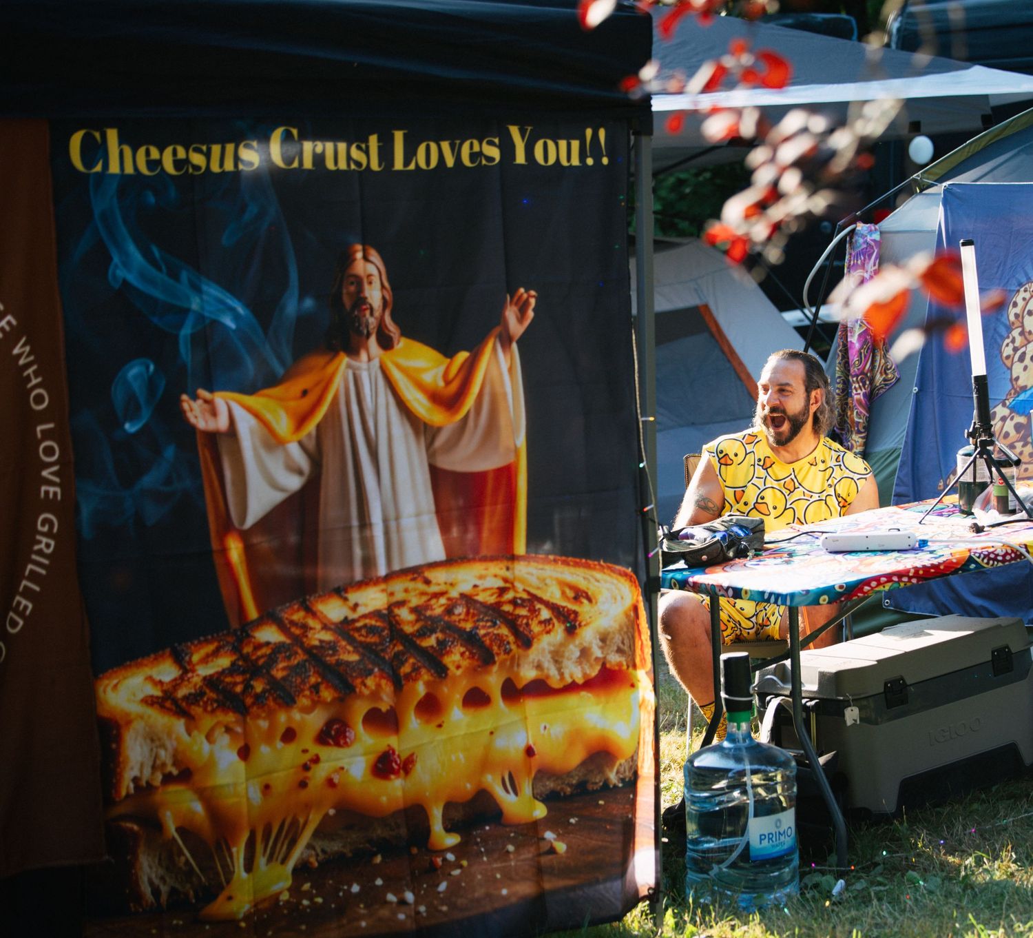 Outdoor food festival booth with humorous Cheesus Crust grilled cheese sign and vendor working at table.