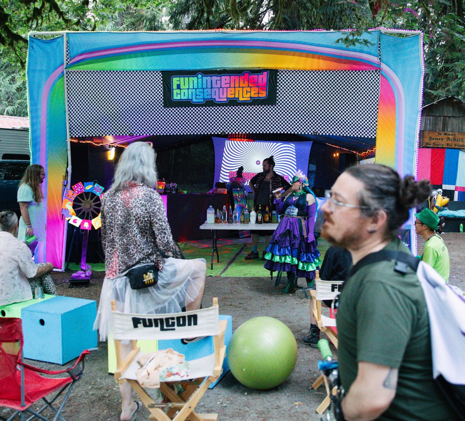 Colorful outdoor festival stage with rainbow LED lights, performers, and audience members seated in director chairs.