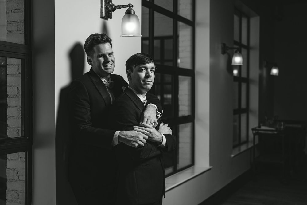 Two grooms embrace and smile in a black and white wedding photo taken in an elegant indoor hallway.