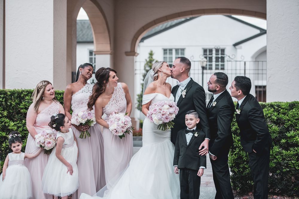 Wedding party poses outdoors as bride and groom kiss, flanked by bridesmaids in blush gowns and groomsmen in black tuxedos.
