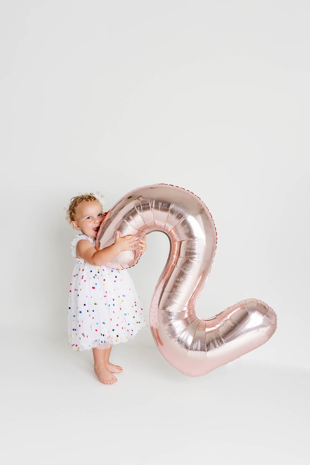 Toddler photoshoot by affordable Riverside county photographer; hugging a rose gold number 2 balloon on white background.