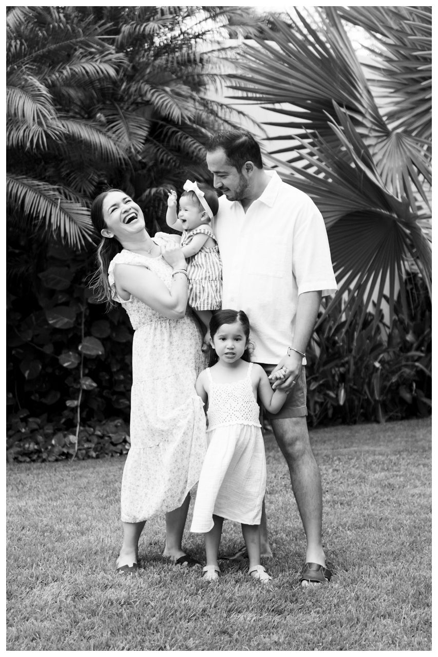 Sayulita family photographer, black and white family portrait of parents holding two young daughters near tropical palm trees outdoors.