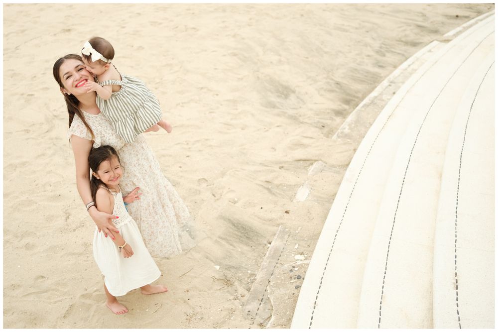 Sayulita family photography, mother smiling with two young daughters on a sandy beach near a white umbrella by the ocean.