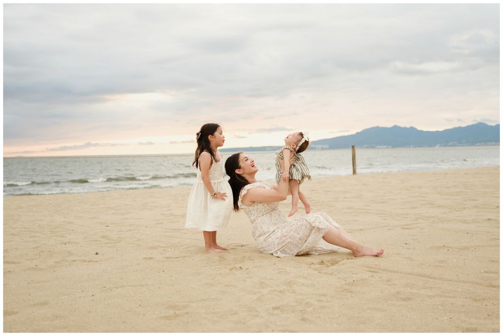 Punta Mita family photography, mother sitting on sandy beach at sunset with two young daughters by the ocean and mountains.