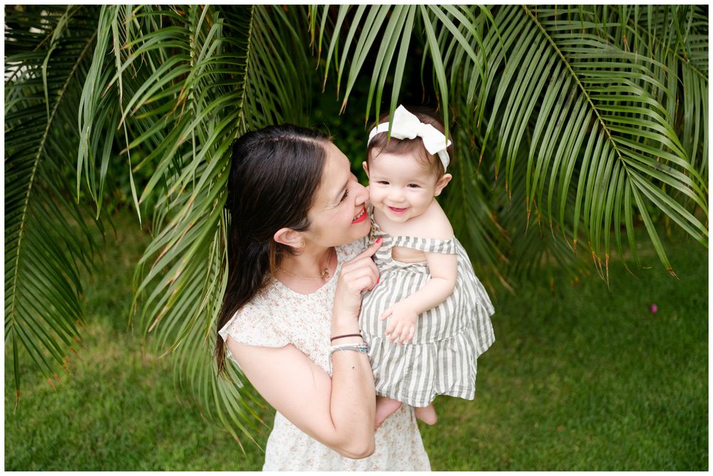 Riviera Nayarit family photography, mother holding her smiling baby girl in front of lush green palm leaves outdoors.