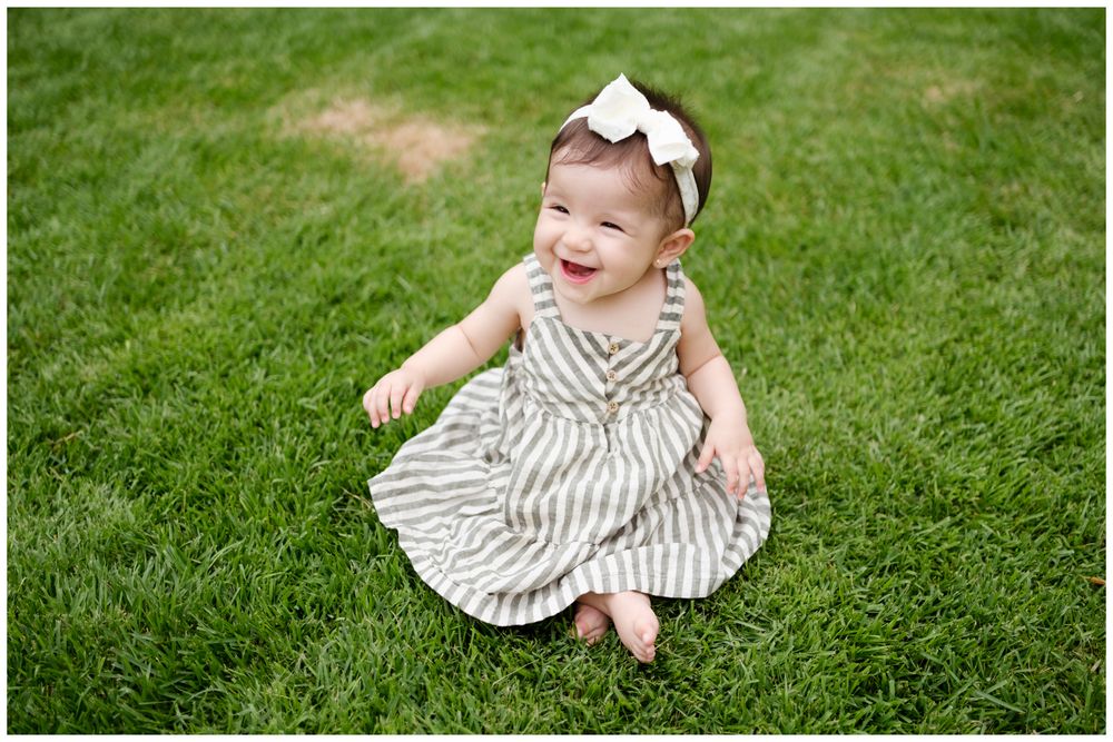 Punta Mita family photography, smiling baby girl in striped dress and white bow headband sitting on lush green grass outdoors.