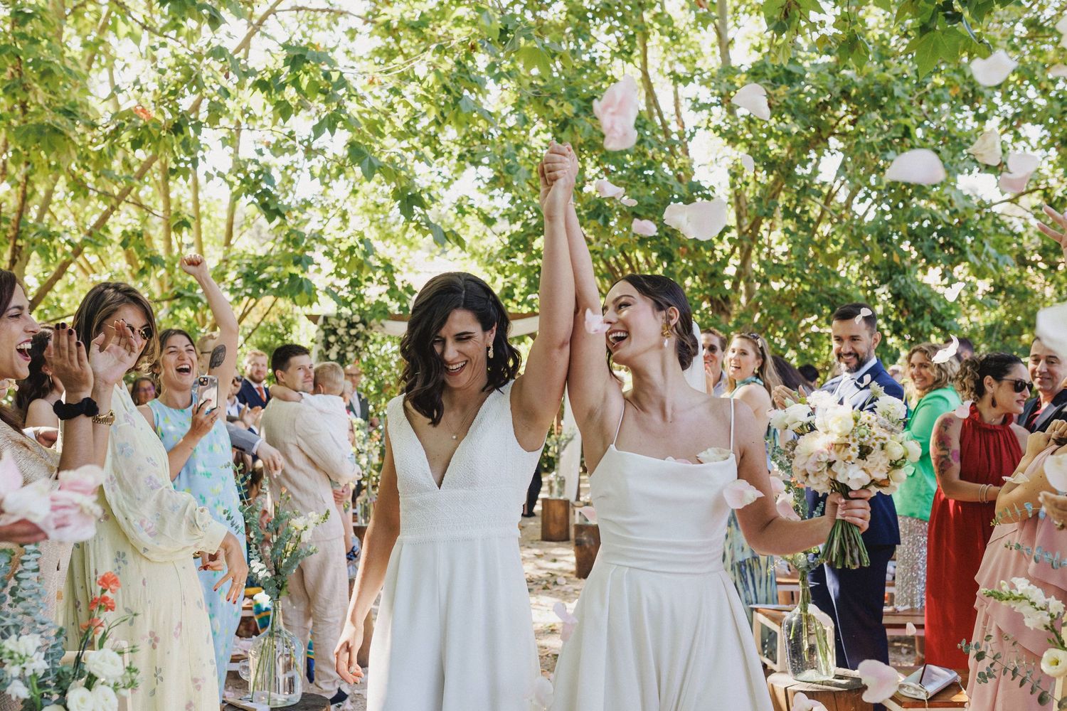 Two people in white dresses hold hands and celebrate during an outdoor garden wedding ceremony