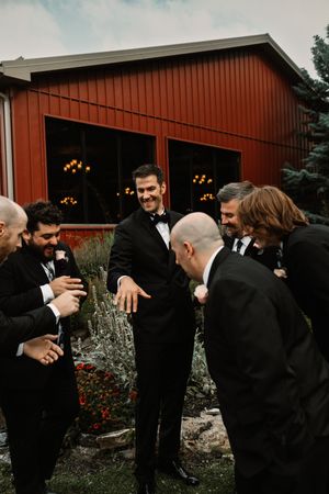 Groom in black tuxedo laughing with groomsmen outside a red barn venue at an elegant wedding celebration.