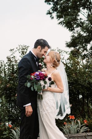 Bride and groom share a romantic nose-to-nose moment outdoors, bride holding a vibrant purple and red bouquet in a lace gown.
