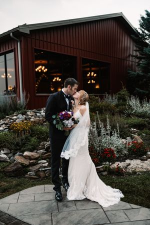 Bride and groom share a romantic kiss outside a red barn venue surrounded by a lush garden at dusk.