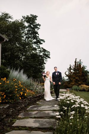 A smiling bride and groom walk hand-in-hand along a stone path surrounded by lush garden flowers at their wedding.