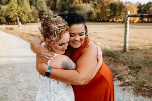 Bride in white lace gown hugging bridesmaid in rust orange dress outdoors at rustic farm wedding venue near Knoxville, TN.