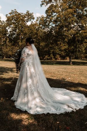 Bride in lace wedding gown with long train and veil stands in sunlit park surrounded by autumn trees near Knoxville, TN.