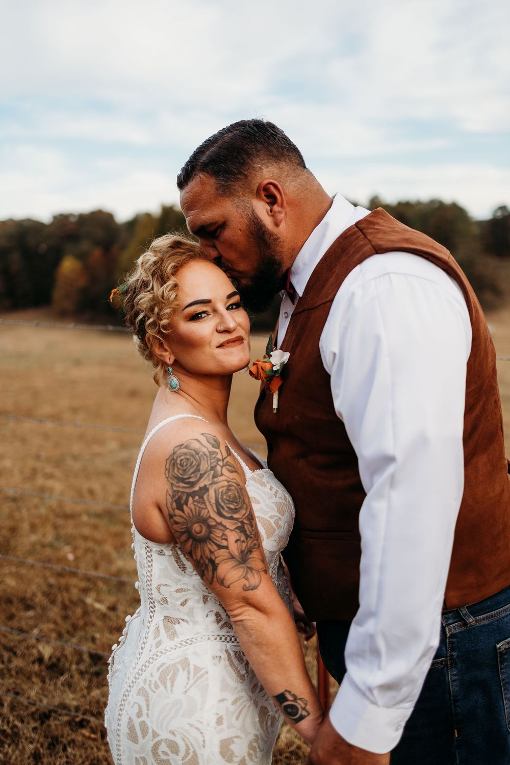 Bride with rose tattoos in lace dress and groom in brown vest share a tender moment in an autumn field near Knoxville, TN.