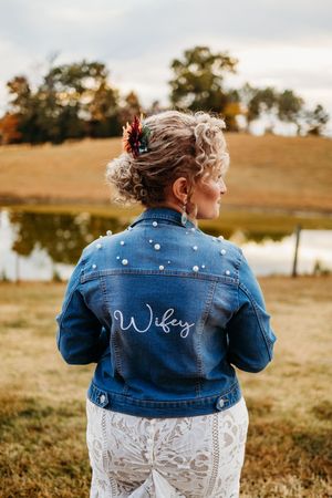 Bride wearing a pearl-embellished denim jacket with Wifey script, lace dress, near a pond in autumn near Knoxville, TN.