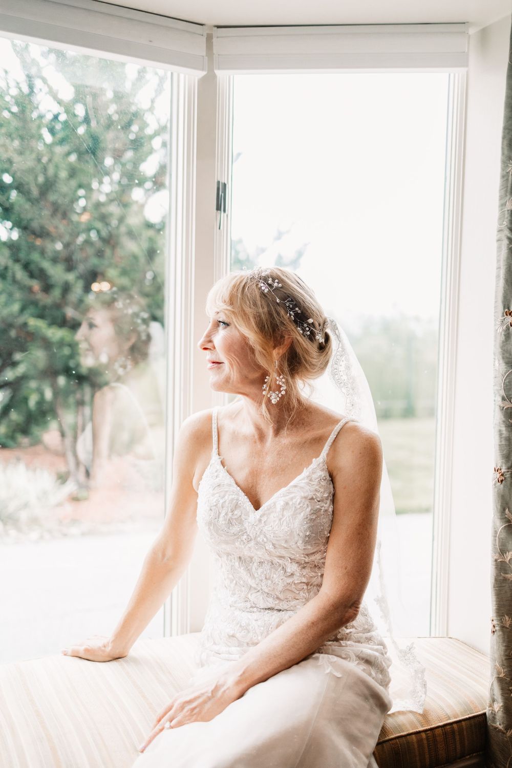 A radiant bride in a lace gown sits by a sunlit window, wearing a veil and floral hair accessories on her wedding day.