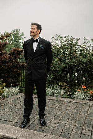 Smiling groom in black tuxedo with bow tie and blue pocket square standing on stone patio surrounded by lush garden greenery.