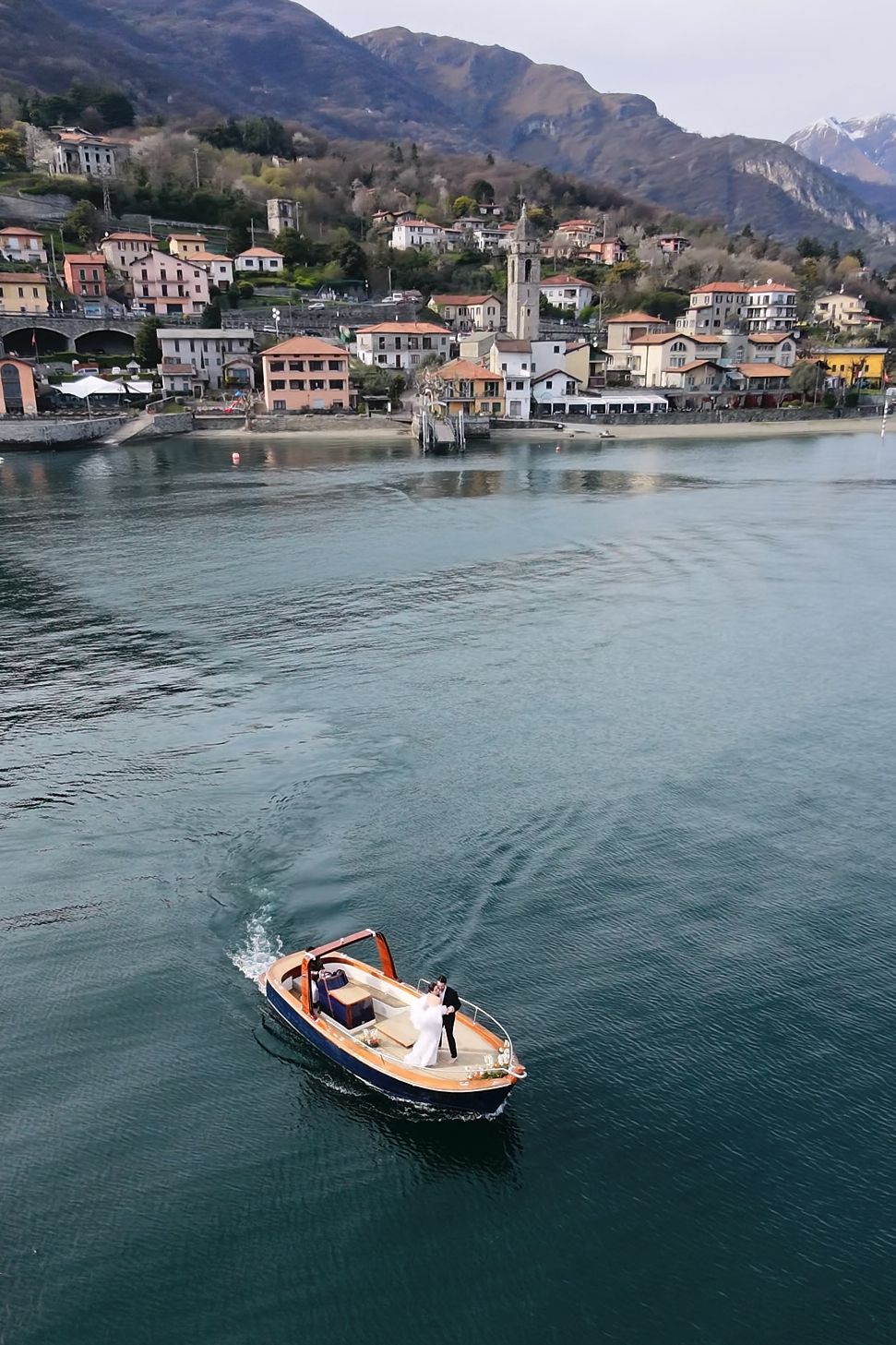 Aerial view of a wooden boat with newlyweds on a turquoise lake with an Italian village and mountains behind.