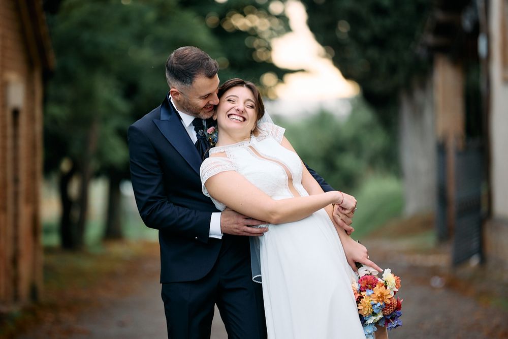 Smiling bride holding colorful bouquet embraced by groom in navy suit outdoors at golden hour.