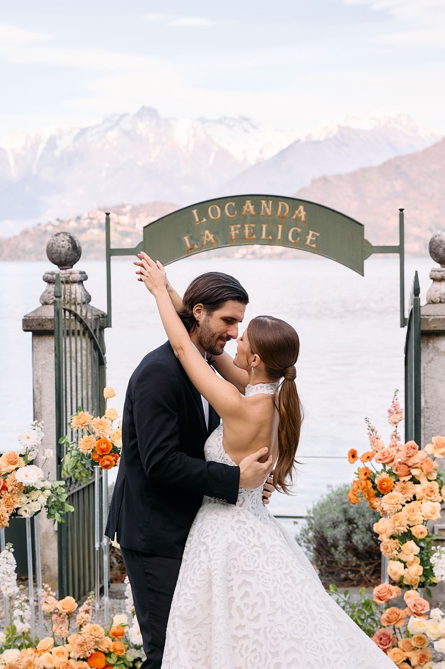 Couple embracing under Locanda La Felice sign surrounded by orange floral arrangements with mountain backdrop.