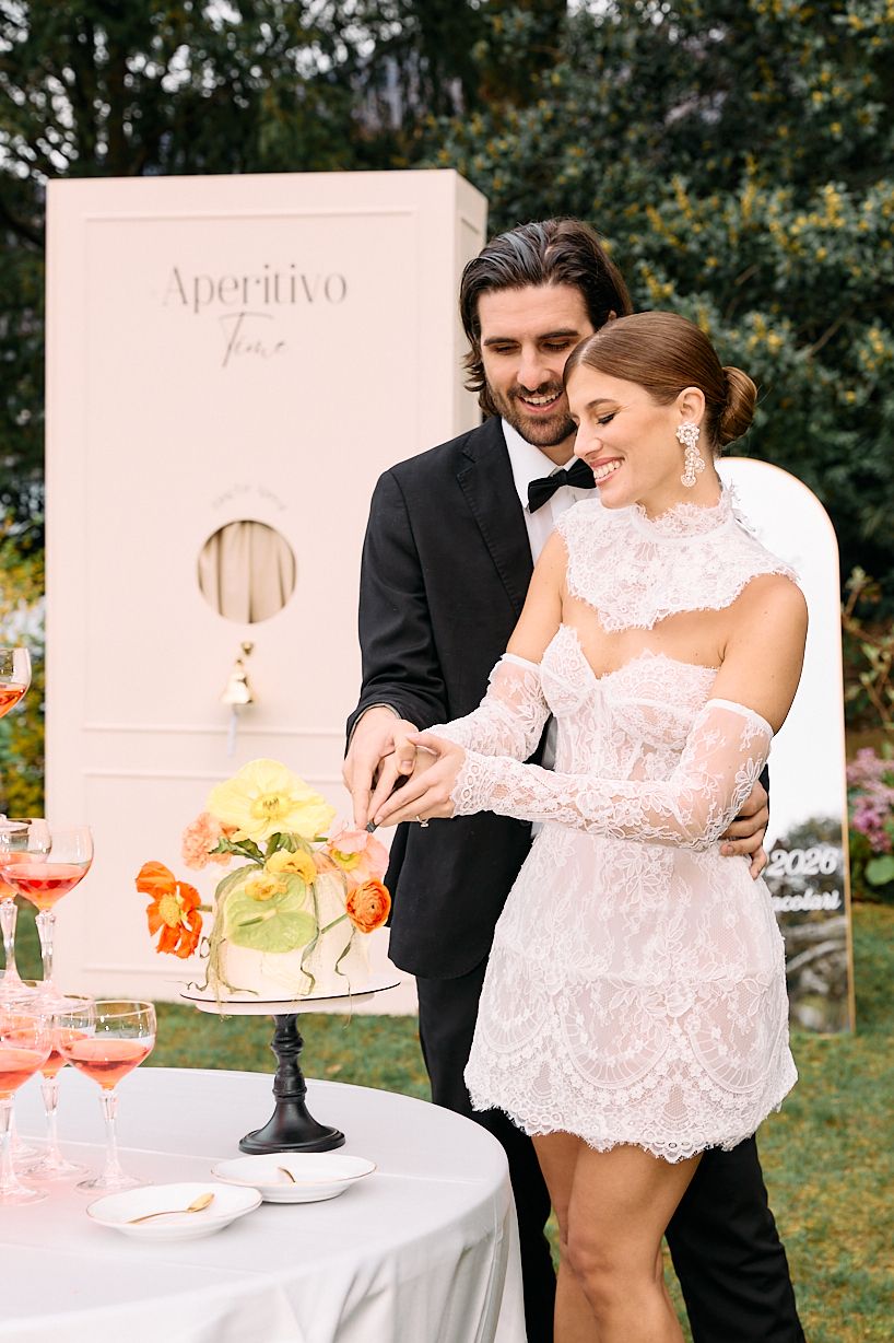Smiling couple in elegant attire cutting a floral wedding cake at outdoor Aperitivo reception.