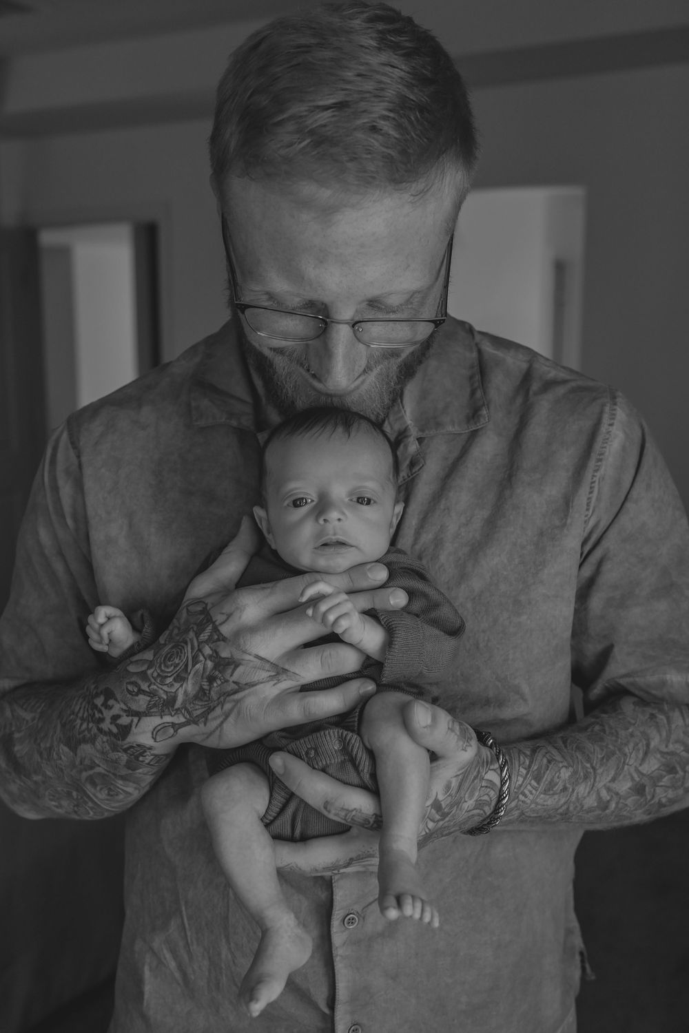 Black and white photo of tattooed man with glasses tenderly holding and kissing a newborn baby.