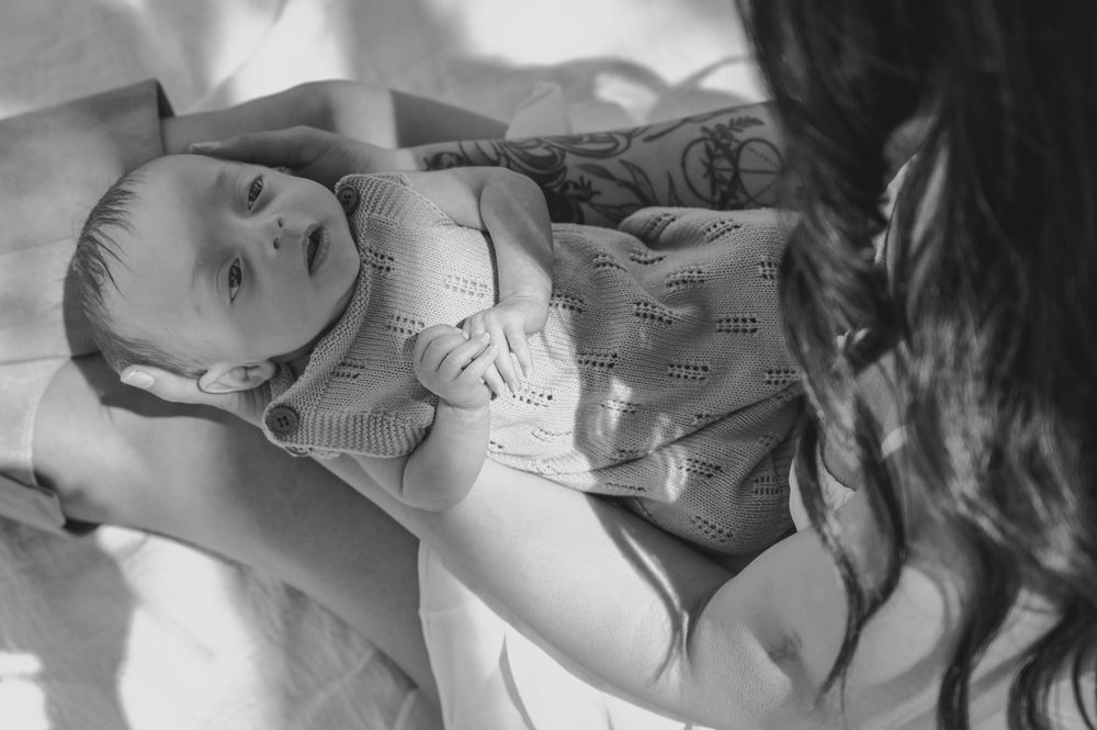 Black and white photo of a newborn baby being cradled by a tattooed mother in soft natural light.