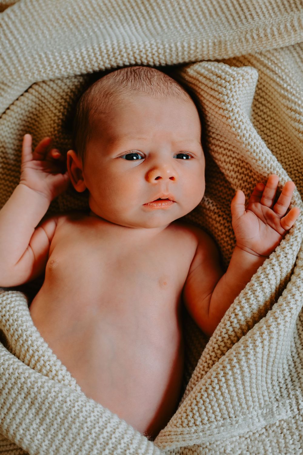 Newborn baby lying on a soft cream knitted blanket, looking up with wide eyes and tiny hands raised.