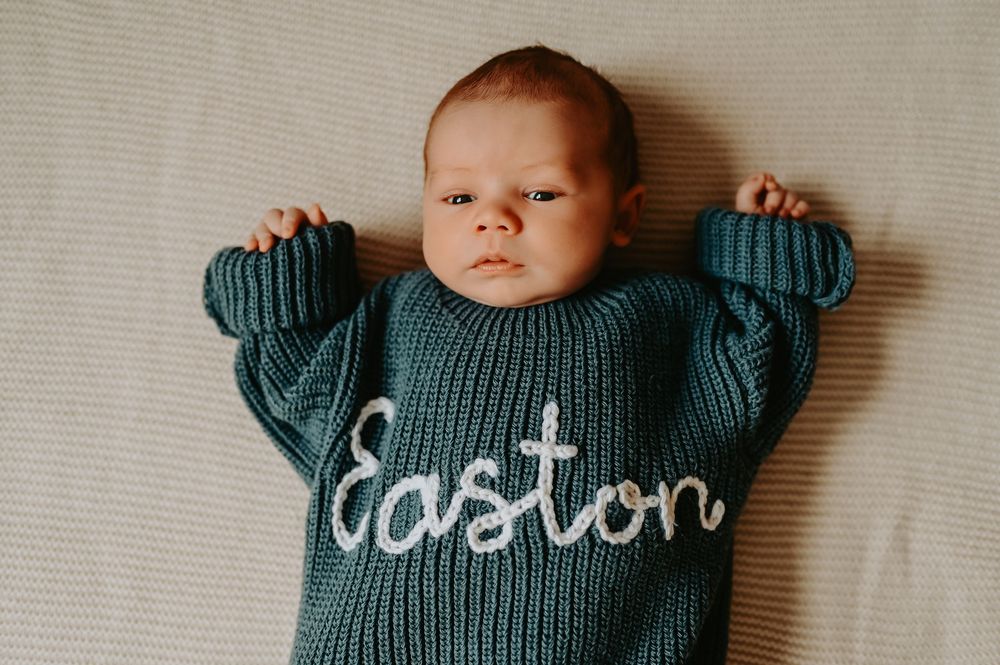 Baby wearing an oversized teal knit sweater with the name Easton embroidered in white cursive script.