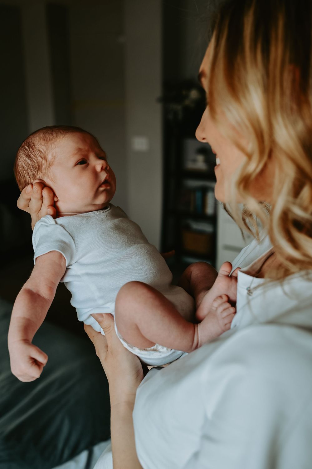 A smiling blonde mother holds her newborn baby in a white onesie, gazing lovingly at the infant.