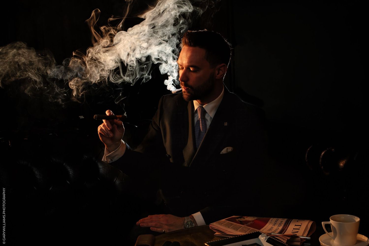 Man in dark suit smoking a cigar at a table, surrounded by dramatic smoke clouds in moody lighting.